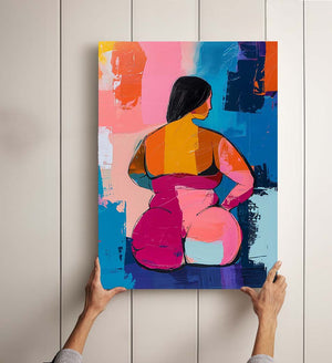 Tableau coloré tenu à deux mains, représentant une femme de dos aux formes généreuses, dans un style peinture au couteau. L’œuvre mêle des aplats vibrants de rose, orange, bleu et jaune, créant une composition expressive et moderne. Fond texturé aux couleurs vives évoquant l’art contemporain et la célébration des corps.