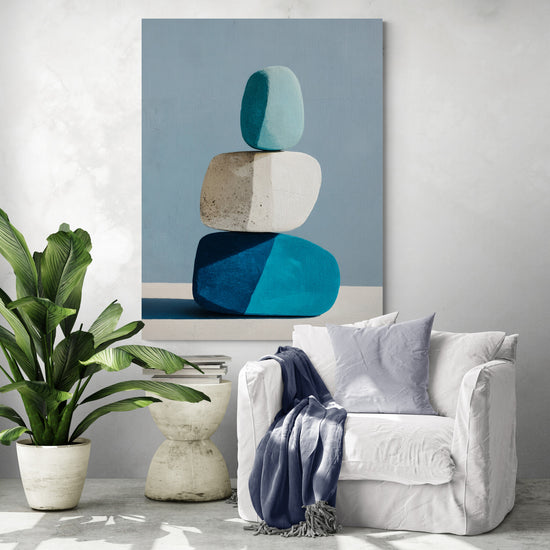 Tableau design aux tons bleus et blancs représentant trois galets empilés, accroché au mur près d’un fauteuil blanc décoré de coussins et plaid bleu, avec une grande plante verte dans un coin lecture lumineux.