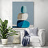 Tableau design aux tons bleus et blancs représentant trois galets empilés, accroché au mur près d’un fauteuil blanc décoré de coussins et plaid bleu, avec une grande plante verte dans un coin lecture lumineux.
