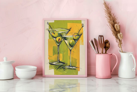 Affiche décorative de deux verres à martini avec olives vertes, encadrée en rose et posée sur un plan de travail de cuisine moderne.