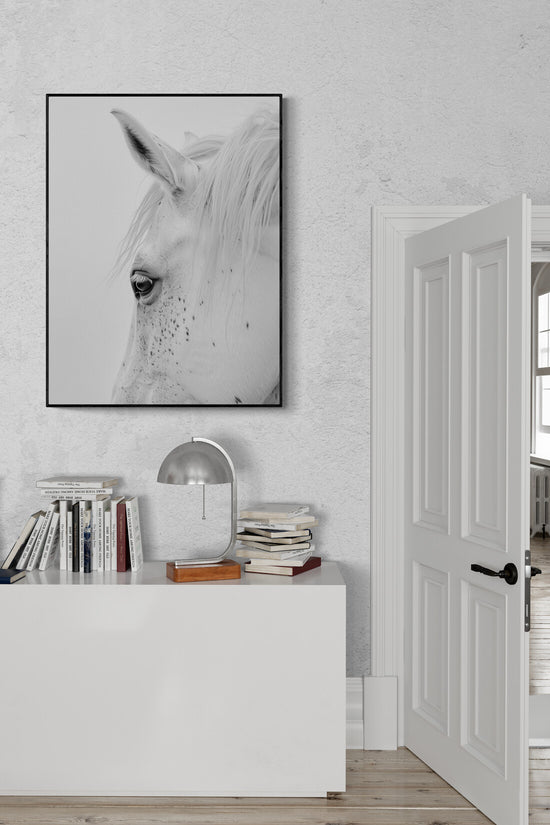 commode blanche, livres empilés, lampe moderne originale, tableau tête de cheval cadre, mur blanc texturé, porte avec moulure blanche.