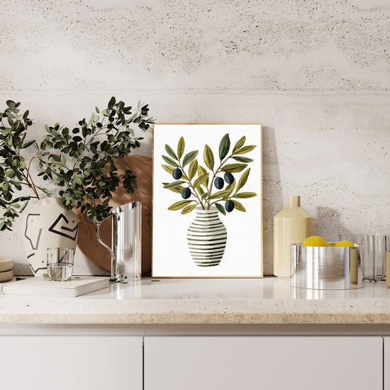 Gros plan du tableau avec un vase rayé noir et blanc contenant des branches d’olivier aux feuilles vertes et aux olives noires, sur fond blanc épuré.