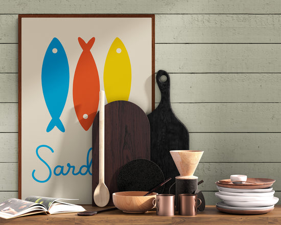 Affiche murale représentant trois sardines stylisées bleu, rouge et jaune, posée sur un plan de travail en bois dans une cuisine rustique, entourée d’ustensiles, planches à découper et vaisselle.