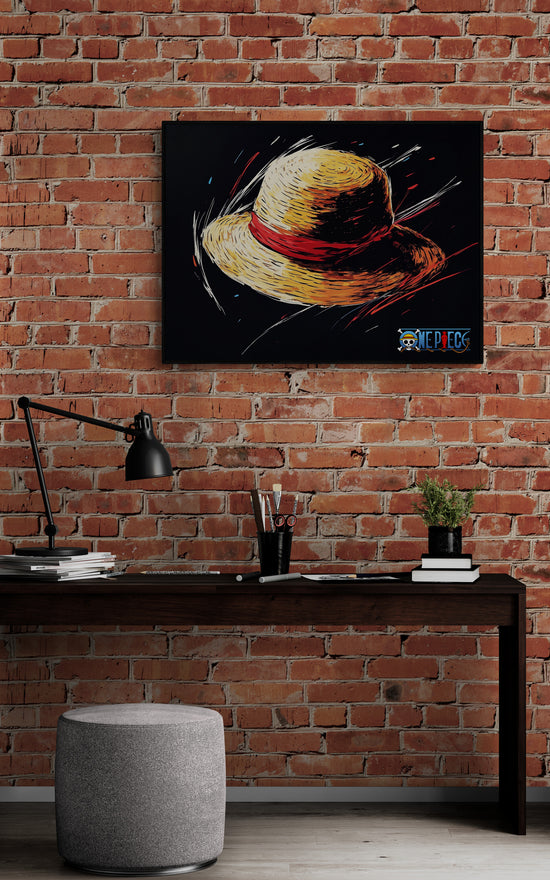 Une toile murale représentant un chapeau de paille avec un ruban rouge sur fond noir. Le cadre est accroché à un mur de briques, au-dessus d’un bureau en bois sombre. Le bureau est décoré avec une lampe articulée noire, des fournitures artistiques et un tabouret gris.