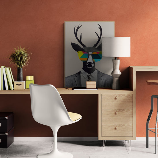 Bureau en bois clair contre un mur terracotta. Une chaise blanche avec coussin jaune fait face au bureau. Le tableau du cerf en costume, posé sur le meuble, ajoute une note décalée. Lampe moderne, fournitures et décoration végétale complètent l’espace.