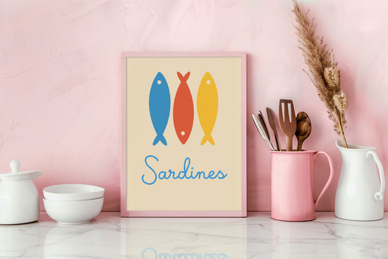 Cadre déco posé sur un plan de travail rose dans une cuisine moderne, illustrant trois sardines stylisées bleu, rouge et jaune avec le mot "Sardines", apportant une ambiance joyeuse et colorée.