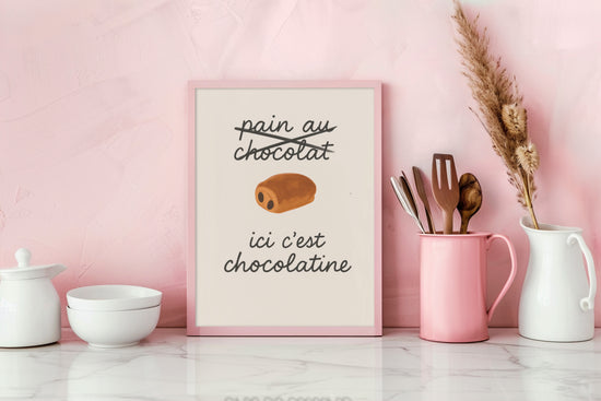Cadre décoratif humoristique avec l’inscription « ici c’est chocolatine », posé sur un plan de travail rose avec ustensiles de cuisine et vaisselle blanche.