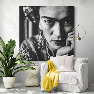 Salon clair avec grand portrait expressif : Affiche géante de Frida Kahlo au mur. Fauteuil blanc, plaid jaune, coussin clair. À gauche, grande plante verte, ambiance paisible et végétale.