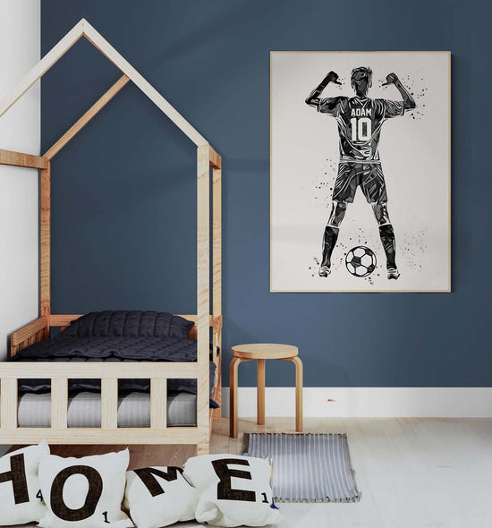 une chambre d'enfant décorée de manière moderne et épurée. Sur le mur, un grand tableau personnalisé représente un joueur de football, vu de dos, avec le prénom "Adam" et le numéro 10 sur son maillot. Le joueur lève les bras en signe de victoire. Le lit en bois simple et des coussins portant les lettres du mot "HOME" complètent l'ambiance chaleureuse et sportive de la pièce.