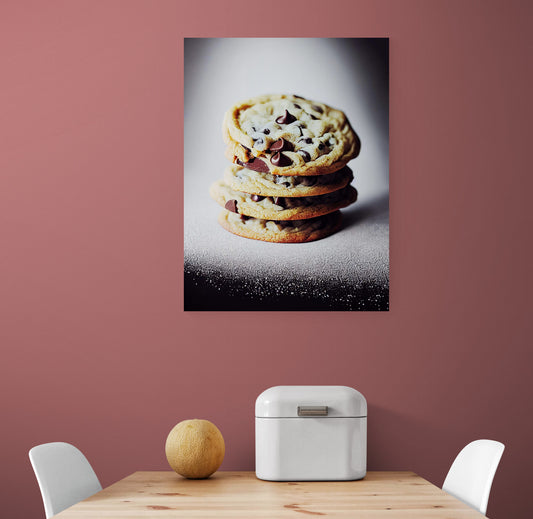 Tableau déco cuisine est accroché sur un mur blanc. C’est une photo de cookie aux pépites de chocolats. Le tableau cuisine est placé ce au-dessus d'une table à manger blanche, deux chaises sont de chaque côté de la table. Une boîte à pain en métal blanc et un melon sont sur la table