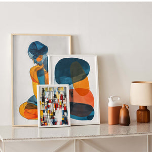 Three framed artistic wall posters placed on a living room furniture. The first features a stylized female silhouette in blue and orange, the second depicts overlapping abstract shapes in blue, orange, and ochre, and the third showcases a colorful geometric composition like a stained glass. To the right, a beige lamp and two ceramic vases complete the modern and cozy interior decoration.