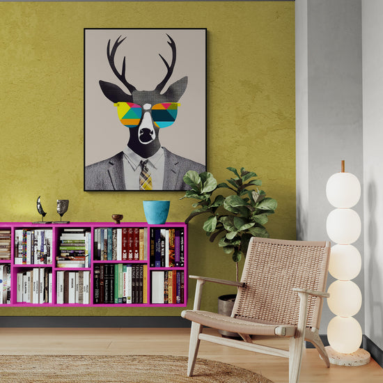 Salon aux tons vifs avec un mur vert olive. Une bibliothèque rose remplie de livres longe le mur. Au-dessus, un tableau de cerf en costume et lunettes colorées apporte une touche pop. Chaise tressée, plante verte et lampe à sphères blanches équilibrent l’ensemble.