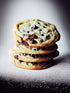 Une photo culinaire avec un zoom sur quatre cookies aux pépites de chocolats posés l'un sur l'autre. Ils sont posés sur une surface noire recouverte de sucre glace