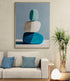 Grand tableau moderne représentant trois galets bleus et blancs empilés, accroché au-dessus d’un canapé beige avec coussins bleus, lampe design et plante verte dans un salon contemporain.