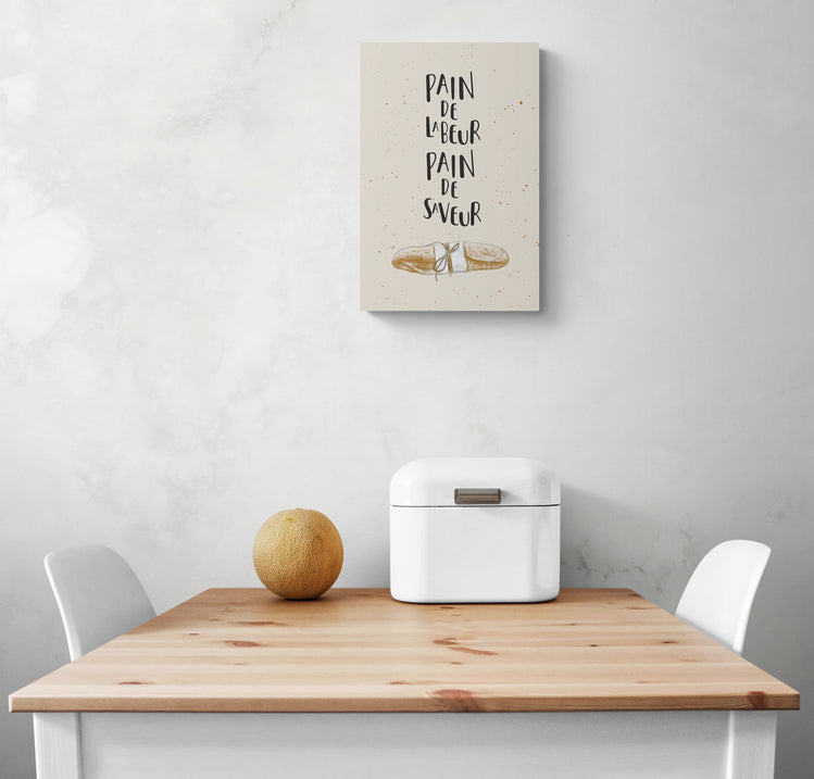 Un tableau aux couleurs neutre est suspendu sur un mur blanc. On peut apercevoir une illustration d'une baguette et une citation. En dessous du tableau, une table en bois et deux chaises blanches à chaque extrémité. Un melon et une corbeille à pain sont posés sur la table
