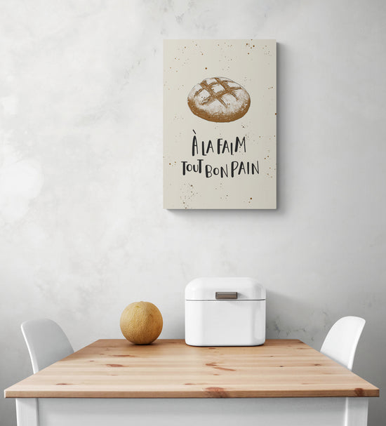 Une toile beige avec une illustration de pain de campagne et une citation est suspendu au mur blanc. Sous le tableau cuisine une table et deux chaises blanches 