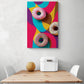 Un tableau coloré de donut avec est accroché sur le mur d'une cuisine blanche. Ce tableau cuisine est placé ce au-dessus d'une table en bois et deux chaises sont de chaque côté. Une boîte à pain en métal blanc et un melon sont sur la table