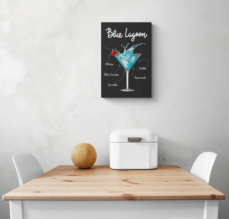 Dans un cadre, une toile en plexiglas avec l'illustration d'un verre de cocktail bleu décore l'intérieur d'une cuisine blanche. Sous le tableau en verre acrylique une table en bois et deux chaises face à face. Une corbeille à pain en métal blanc et un fruit rond sont posés sur la table.