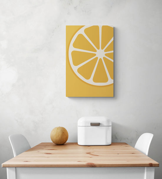 Un tableau pour cuisine est suspendu au centre d'un mur blanc. En dessous du tableau orange se trouve une table et deux chaises. Sur la table se trouve une boite à pain en métal blanc et un petit melon. 