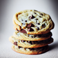 Une photo culinaire avec un zoom sur quatre cookies aux pépites de chocolats posés l'un sur l'autre. Ils sont posés sur une surface noire recouverte de sucre glace