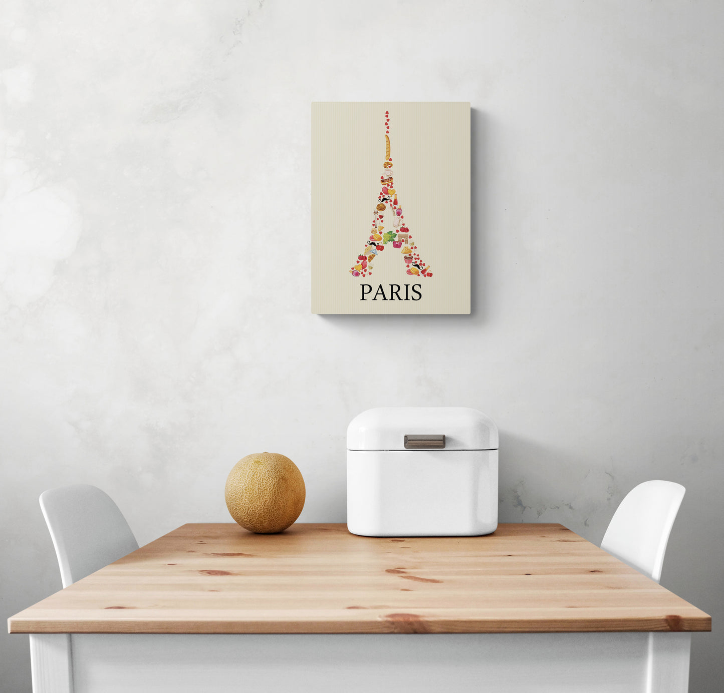 Sur un mur blanc d'une cuisine, un tableau cuisine original aux couleurs douces représentant une image de la Tour Eiffel est accroché. La photo montre également une table en bois et deux chaises blanches qui semblent être disposées de manière à créer un espace accueillant.