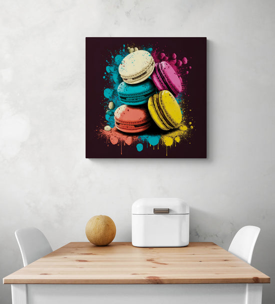 Une toile macaron est disposée au centre de l'image. Le tableau est dans un style pop art avec des éclaboussures sont autour des macarons bleu, rose, orange, jaune et blanc. Le fond du tableau pour cuisine est noir. Dessous une table de repas blanc et bois naturelle aux extrémités deux chaises blanche un melon et une boîte à pain sont sur la table.